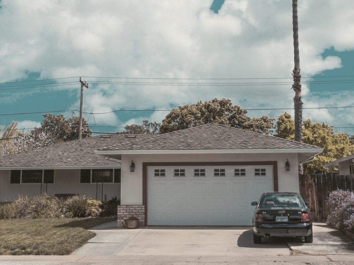 Single-story ranch home with an attached garage, mature trees, and a concrete driveway on a sunny suburban street.